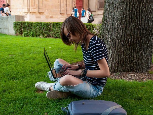 A teenage woman studying with a laptop sitting on the grass in a park of a university in a university campus - Photo, Image