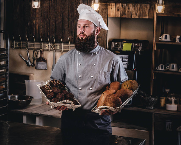 Handsome bearded chef in uniform showing tray of fresh bread in the kitchen of bakery. - Photo, Image