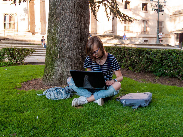 Une adolescente étudiant avec un ordinateur portable assis sur l'herbe dans un parc d'une université dans un campus universitaire
 - Photo, image