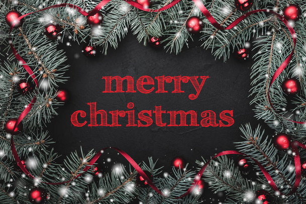 Black stone background with a frame of fir branches decorated with balls and red ribbon. Top view. With Merry Christmas text - Photo, Image