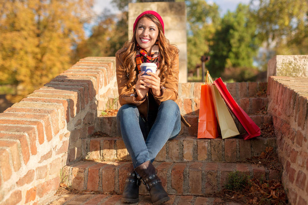 Красивая молодая женщина сидит на лестнице снаружи, выпивая кофе и улыбаясь после покупок
  - Фото, изображение