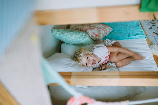 Linda niña rubia sonriendo y acostada en la cama cubierta de almohadas
 - Foto, imagen