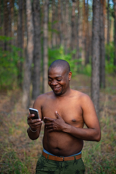 African-American man without a shirt holding a smartphone and laughing in the woods - Foto, afbeelding
