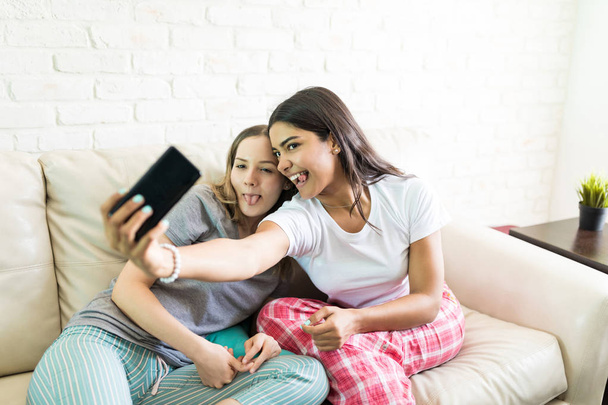 Funny females taking selfie while sticking out tongue via mobile phone at home - Photo, Image
