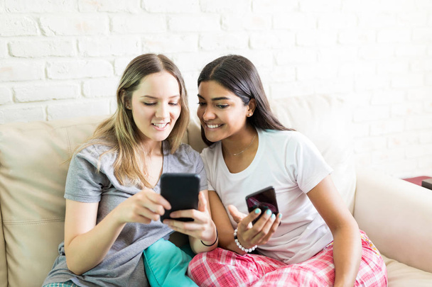 Lächelnde Freundinnen, die Dateien vom Smartphone auf ein anderes übertragen, während sie zu Hause auf dem Sofa sitzen - Foto, Bild