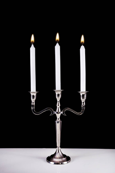 silberner Barockkerzenständer mit drei weißen Kerzen aus Paraffin, die auf dem Schreibtisch brennen und schwarzem Hintergrund. romantisches und stilvolles Konzept - Foto, Bild