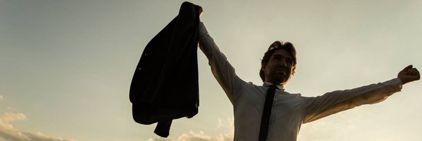 Biznesmen zwycięsko stojąc z rękami podniósł wysoko trzymając jego kurtka w jednym przed wieczornym niebie. - Zdjęcie, obraz