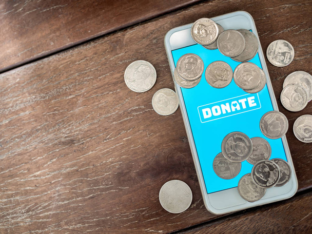 flache Lage spenden Symbol-Taste auf blauem Bildschirm auf weißem Handy mit Münzen thailändischen Baht Geld auf hölzernem Hintergrund mit Kopierraum. Online-Spendenkonzept. - Foto, Bild