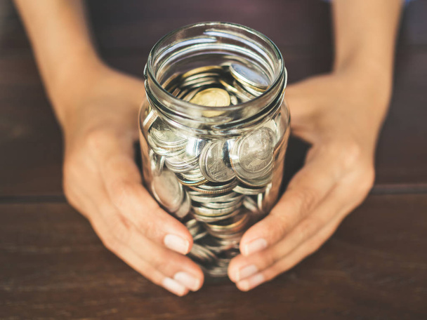 Coins in glass jar in hands on wooden table background. Saving money and donation concept. - Photo, Image