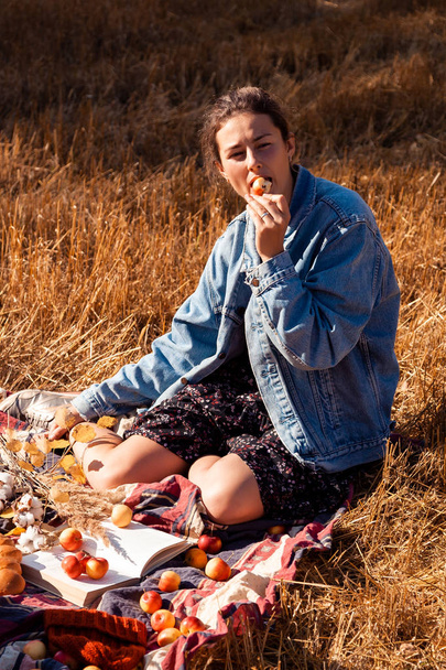 Piknik a friss levegőn: egy fiatal nő, egy farmer kabát és ruha eszik almát, és élvezi természet, ül egy kockás egy piknik kosár, alma, bor. A háton és a háttérben van a mező őszi táj. - Fotó, kép