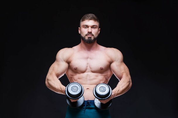 Muscular bodybuilder guy doing exercises with dumbbell over black background - Фото, изображение