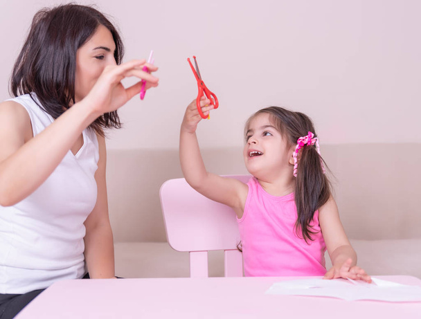 La madre le enseña a su hija a usar tijeras y papel cortado mientras está sentada en la mesa rosa. Enfoque selectivo y pequeña profundidad de campo
. - Foto, Imagen