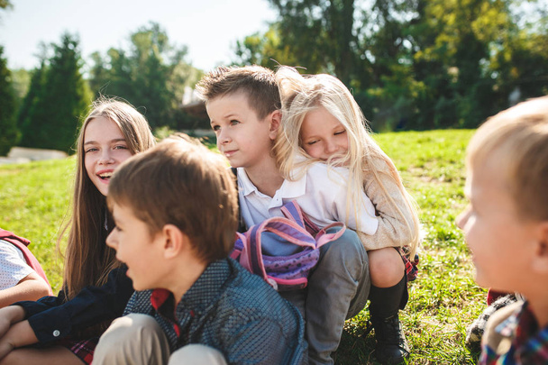 Ryhmä koulun ja esikoulun ikäisiä lapsia istuu puistossa vihreällä ruoholla.
. - Valokuva, kuva