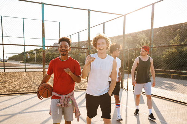 Photo of young sporty men smiling and holding ball while standing at basketball playground outdoor during summer sunny day - Fotoğraf, Görsel