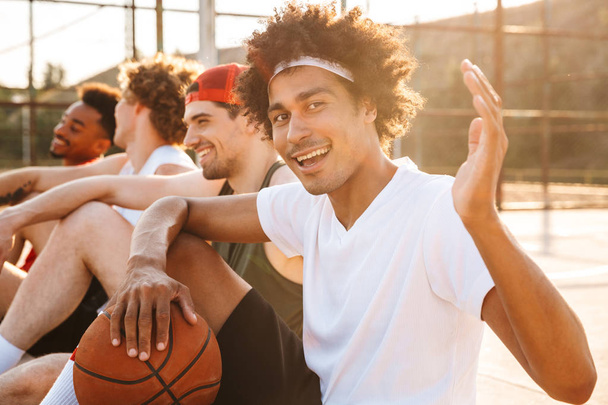 Group of cheerful multiethnic friends basketball players sitting at the sports ground, talking, greeting - Fotoğraf, Görsel