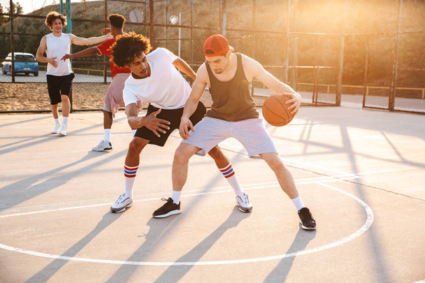 Ryhmä nuoria monikansallisia miehiä koripalloilijoita pelaamassa koripalloa urheilukentällä
 - Valokuva, kuva
