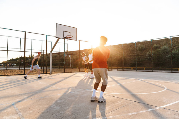 Grupo de jóvenes y alegres jugadores multiétnicos de baloncesto jugando al baloncesto en el campo de deportes, puesta de sol
 - Foto, imagen