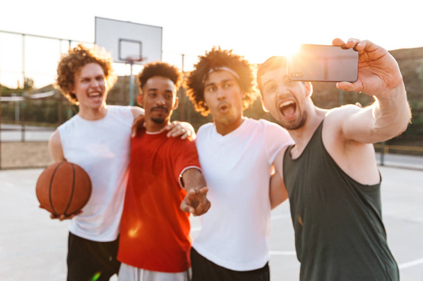 Portrait of cheerful sporty men smiling and taking selfie on smartphone while playing basketball at playground outdoor during summer sunny day - Photo, image