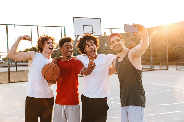 Strong multiethnic men smiling and taking selfie on smartphone while playing basketball at playground outdoor during summer sunny day - Φωτογραφία, εικόνα