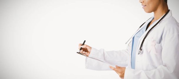 Female doctor wearing breast cancer awareness pink ribbon writing on a note pad on a white background - Foto, Bild