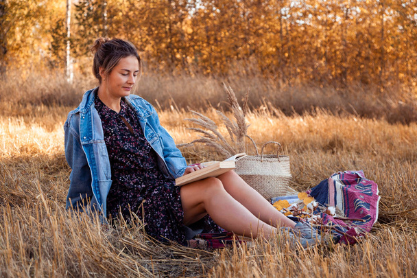 The concept of livestyle and family outdoor recreation in autumn. Picnic in the fresh air: a young woman in a denim jacket and dress read books on plaid with a picnic basket, apples, wine. On the  background autumn  field. - Photo, Image