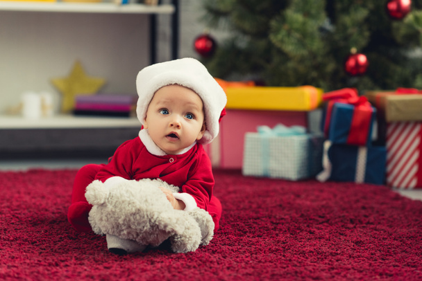 close-up portrait of adorable little baby in santa suit lying on red carpet with teddy bear in front of christmas tree and gifts - Фото, изображение