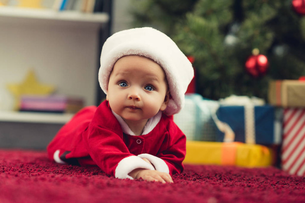 close-up portrait of beautiful little baby in santa suit lying on red carpet in front of christmas tree and gifts - Photo, Image