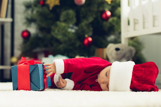 adorable little baby in santa suit lying on floor with christmas gift box - Фото, изображение