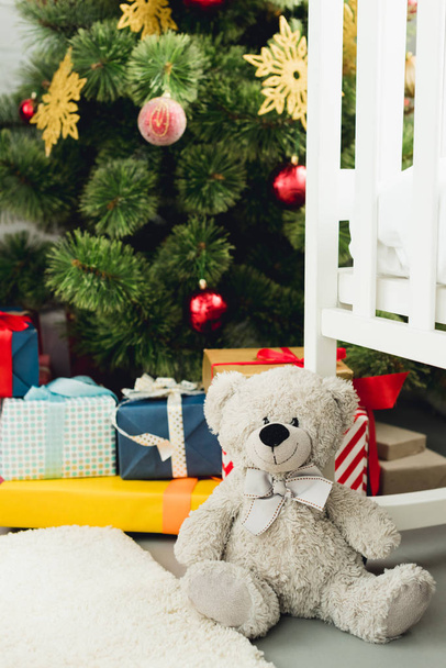 oso de peluche apoyado en la cuna delante del árbol de Navidad y regalos
 - Foto, Imagen