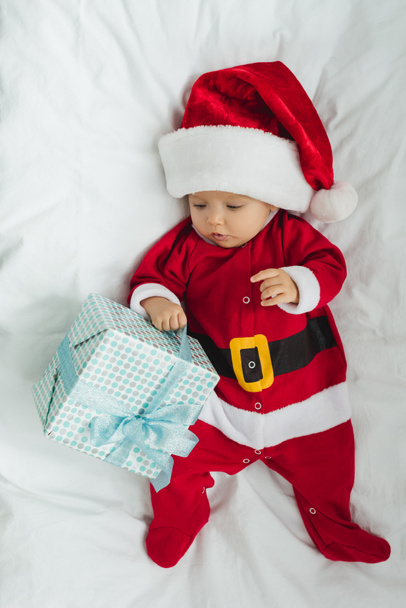 Blick von oben auf entzückendes kleines Baby im Weihnachtsmann-Anzug in der Krippe mit Weihnachtsgeschenk - Foto, Bild