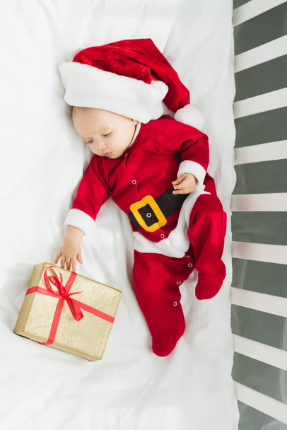 vista superior de hermoso bebé en traje de santa acostado en cuna con regalo de Navidad
 - Foto, Imagen