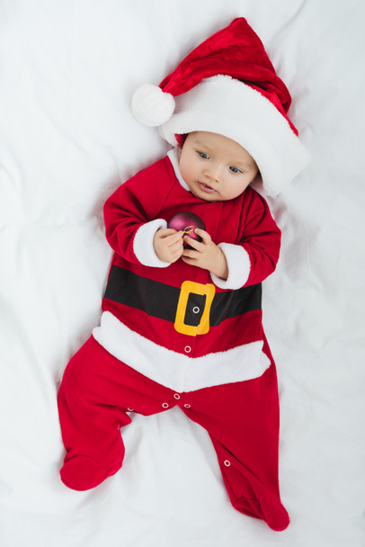 vista superior de adorable bebé en traje de santa celebración de la navidad bauble mientras está acostado en la cuna
 - Foto, Imagen