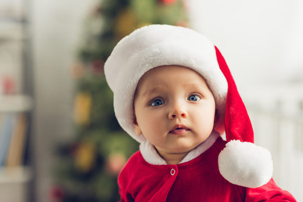 close-up retrato de adorável bebê em santa chapéu olhando para a câmera com árvore de natal borrada no fundo
 - Foto, Imagem