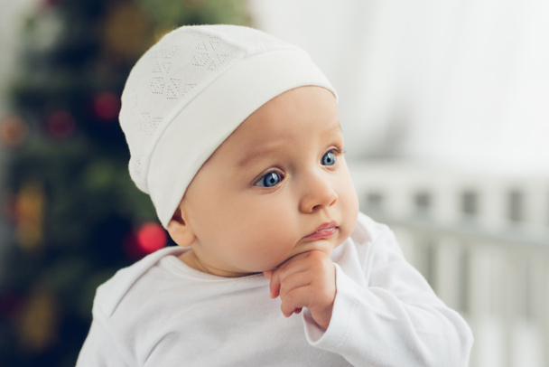 close-up portrait of cute little baby in white hat with blurred christmas tree on background - Photo, Image