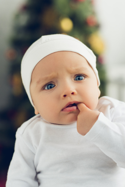close-up portrait of confused little baby in white hat with blurred christmas tree on background - Photo, Image