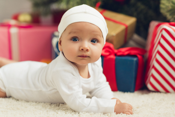 szczegół portret mało noworodek leżąc na podłodze z Boże Narodzenie prezenty niewyraźne na tle i patrząc na kamery - Zdjęcie, obraz