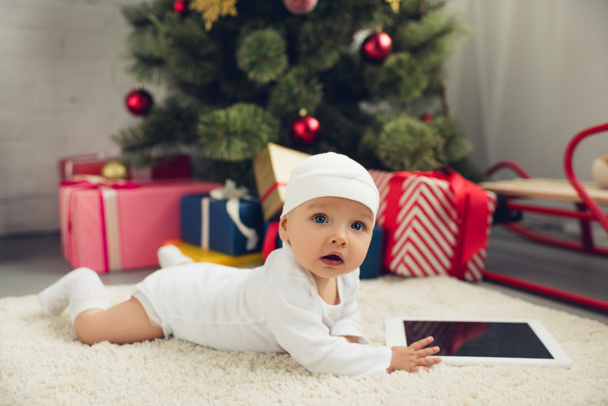 cute little baby with tablet lying on floor with christmas gifts and tree blurred on background - Fotoğraf, Görsel