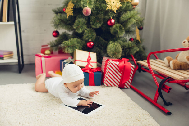 adorable little baby with tablet lying on floor near christmas gifts and tree - 写真・画像