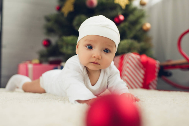entzückendes kleines Baby auf dem Boden liegend mit Weihnachtsbaum im Hintergrund verschwommen - Foto, Bild
