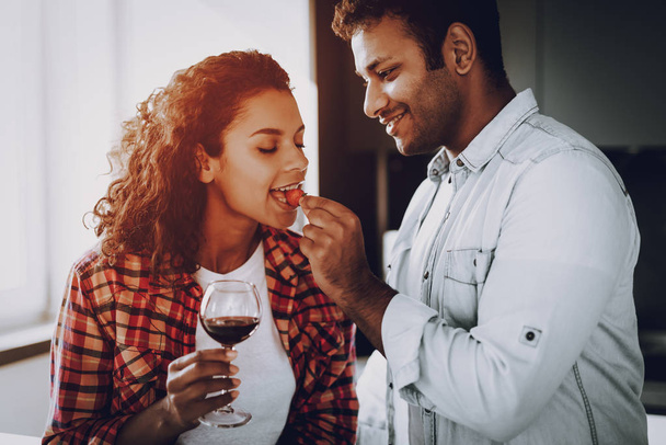 Boyfriend Is Feeding Girlfriend With Strawberry. Weekend Concept. Chilling At Kitchen. Holiday Resting. Smiling Together. Having Fun. Romantic Date. Afro American Couple Drinking Wine. - Photo, Image