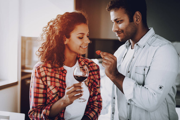 Boyfriend Is Feeding Girlfriend With Strawberry. Weekend Concept. Chilling At Kitchen. Holiday Resting. Smiling Together. Having Fun. Romantic Date. Afro American Couple Drinking Wine. - Fotoğraf, Görsel