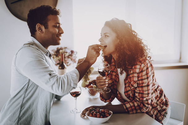 Boyfriend Is Feeding Girlfriend With Strawberry. Weekend Concept. Chilling At Kitchen. Holiday Resting. Smiling Together. Having Fun. Romantic Date. Afro American Couple Drinking Wine. - Foto, imagen