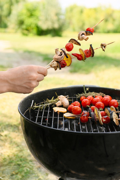 Man holding skewers with vegetables over barbecue grill outdoors - Foto, imagen
