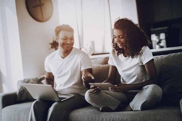 Afro American Couple With Gadgets On The Couch. Morning Leisure. Have A Break. Look On The Screen. Chilling On Sofa. Home Clothes. Holiday Resting. Smiling Together. Laptop And Tablet. - 写真・画像