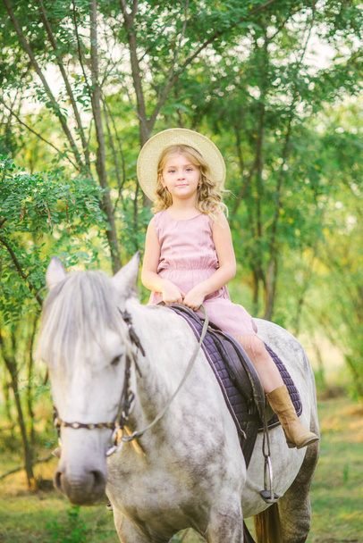 Little cute girl with light curly hair in a straw hat riding a horse at sunset on a sunny warm autumn day - 写真・画像