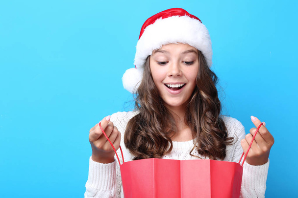 surprised teenage girl in santa hat holding red shopping bag on blue background - Foto, afbeelding