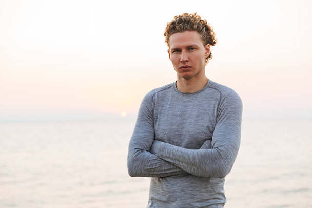Serious Curly Sportsman posing with crossed arms and looking at the camera while being on beach near the sea - Фото, изображение