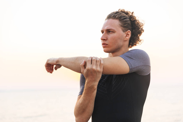 Photo of young handsome sports man outdoors on the beach make stretching exercises. - Φωτογραφία, εικόνα