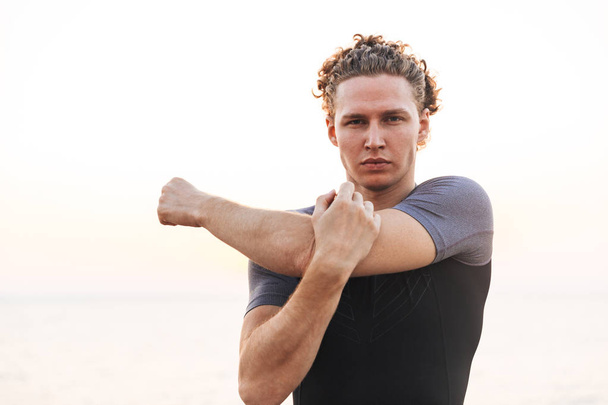 Serious Curly Sportsman warming up and looking at the camera on beach near the sea - Foto, Bild