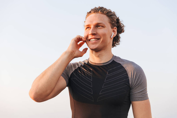 Smiling young sportsman in wireless earphones standing outdoors over blue sky - Фото, изображение
